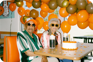 Two elderly people sitting at a table with balloons in the background