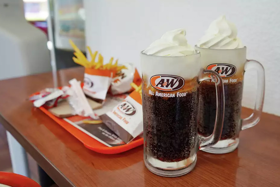 2 Root Beer Floats in A&W Mugs sitting on a table. A tray of A&W food is to the left of the mugs.