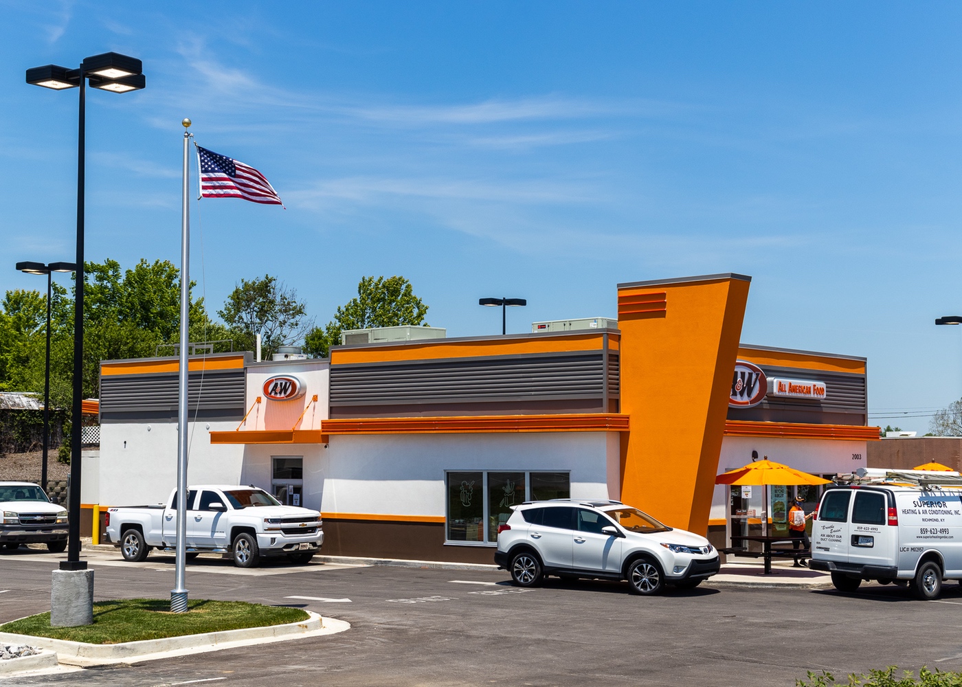 Exterior side view photo of A&W Restaurant showing cars in drive-thru and team member walking bag out to car.