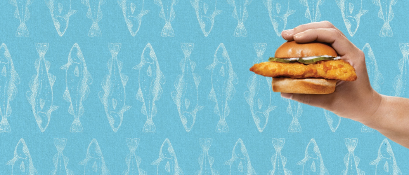 Background is blue with drawings of fish. Person holding Cod Slider in their hand on the right side of image.