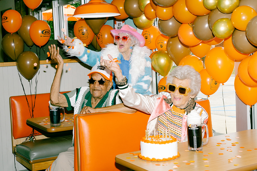 3 people sitting at an orange A&W booth surrounded by orange and gold balloons. An orange and white cake & a Root Beer Float are sitting on the table.