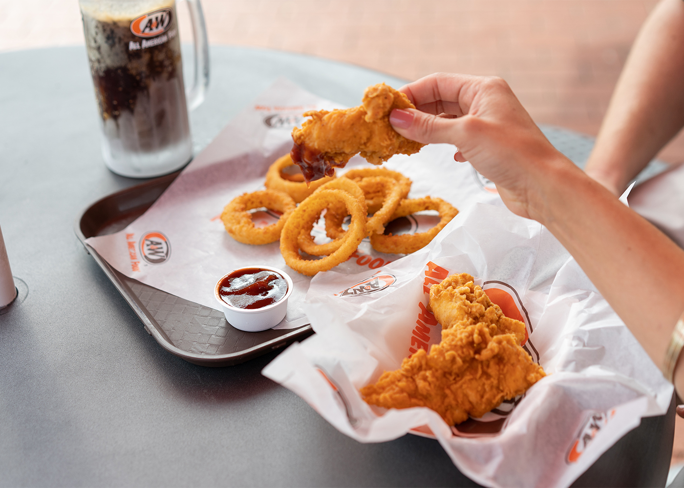 Person holding Chicken Tender. Root Beer and Onion Rings are sitting on a table.
