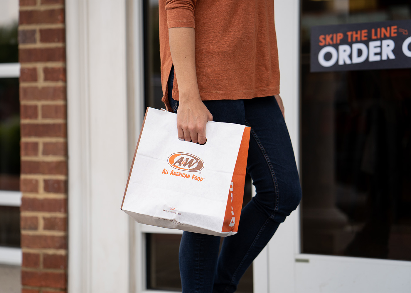 Person is walking outside and holding an A&W Restaurants to-go bag in their right hand.