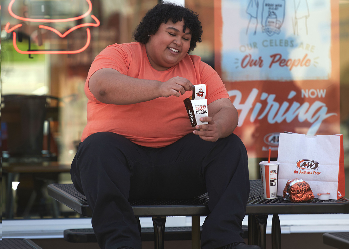 Photo of an A&W Team Member sitting on a table outside the A&W Restaurant. She is holding a box of Cheese Curds and an A&W to-go bag is on the table next to her.