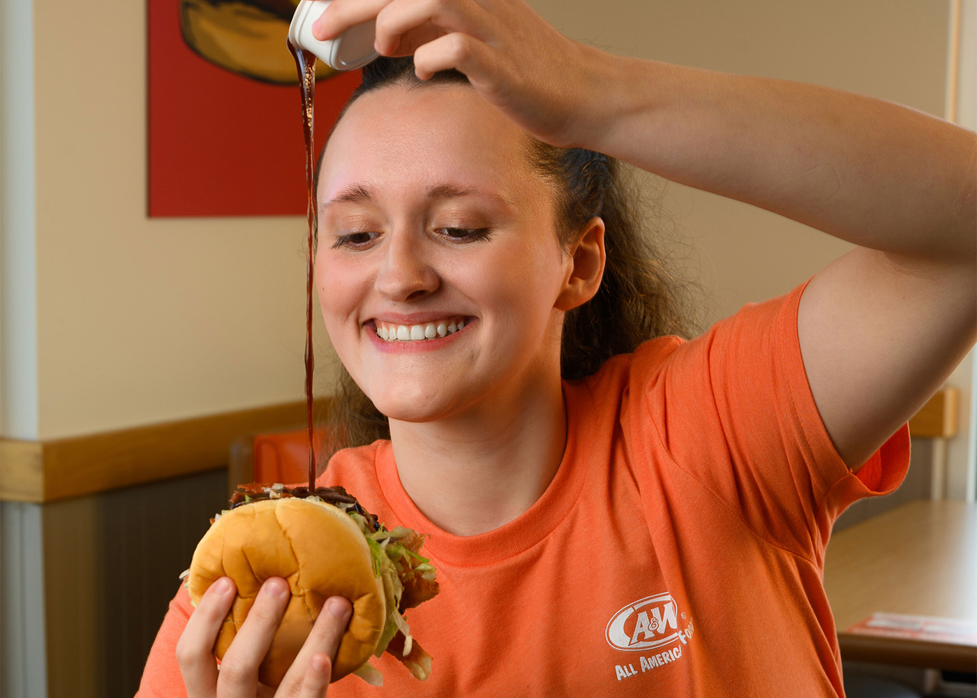 A&W Team Member inside the restaurant. She is holding a burger and drizzling BBQ sauce onto the burger. 