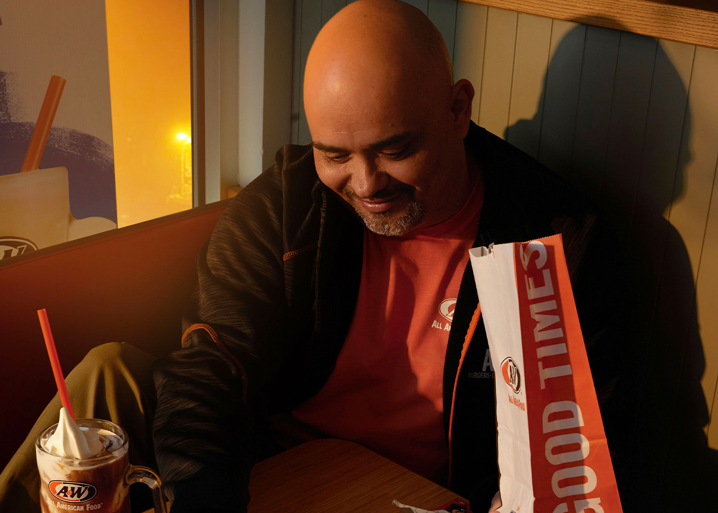 Photo of A&W Team Member sitting at a table inside the restaurant. There is a Root Beer Float on the table and an A&W to-go bag. He is picking up a fry with his right hand.
