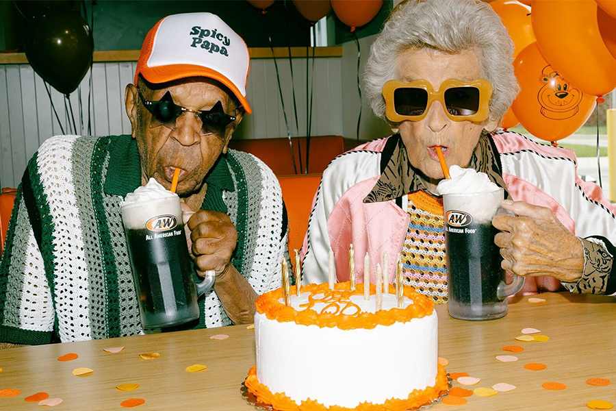 A man and woman sitting at a table drinking Root Beer Floats. An orange and white cake is sitting on the table.