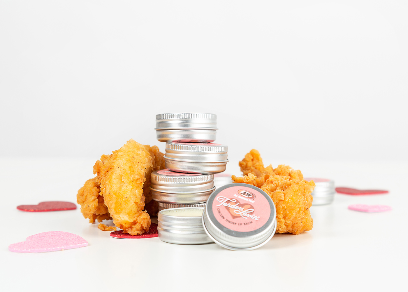 Tins of Tender Lovin' Lip Balm are stacked on top of one another. The tins of Lip Balm are surrounded by Hand-Breaded Chicken Tenders and pink hearts.